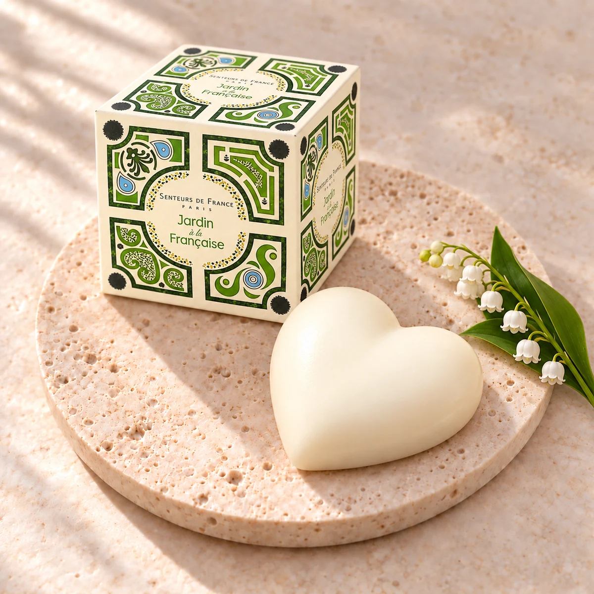 Cremefarbene Muguet-Herzseife von Senteurs de France auf einem Travertinteller mit grün illustrierter Würfelschachtel und weißen Maiglöckchen.