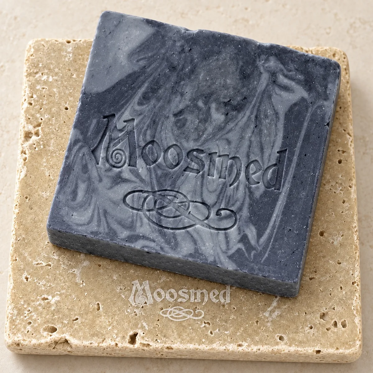 Dunkelgraue Moosmed African Black Soap mit heller Marmorierung und geprägtem Logo auf einem quadratischen Travertinteller.