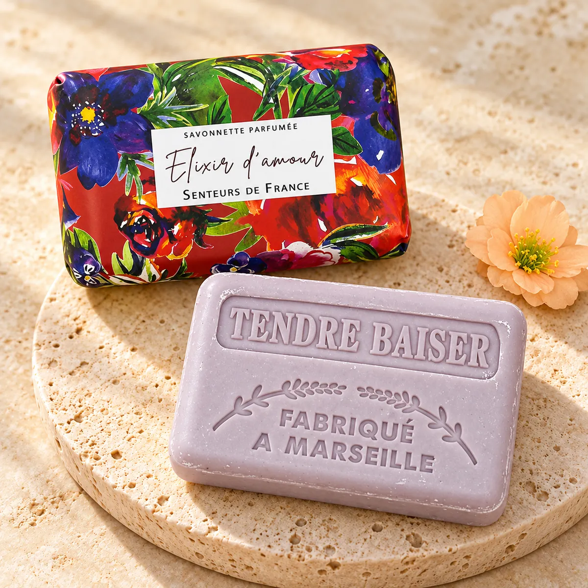 Hellviolette Elixir-d’amour-Seife mit Tendre-baiser-Prägung auf Travertin, mit roter Blumenverpackung und pfirsichfarbener Blüte.