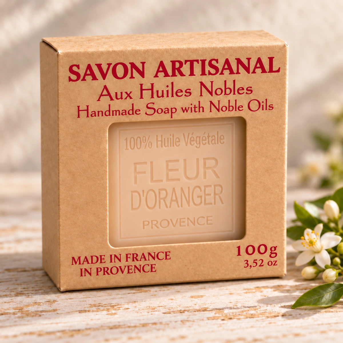 Maître Savonitto Seife Orangenblüte in vollständig sichtbarer Kraftkartonverpackung mit Sichtfenster, neben einem Orangenblütenzweig auf hellem Holz.