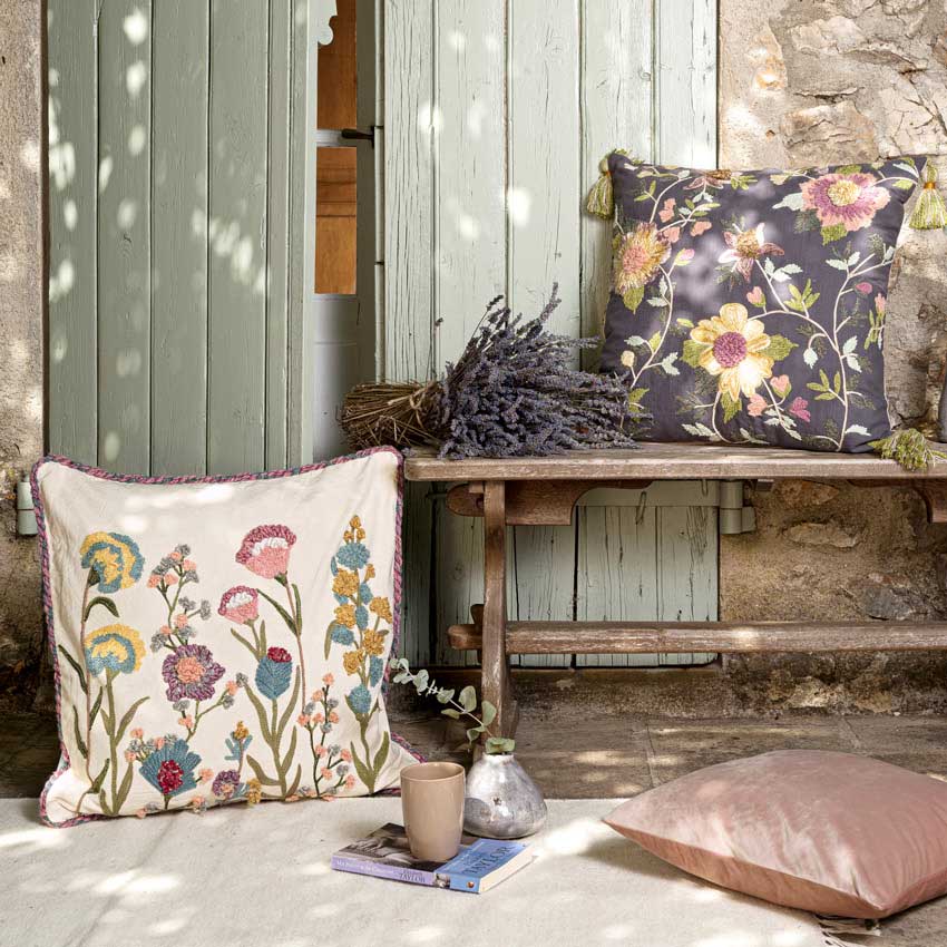 Magma Kissenserie „Amelie“ mit romantischen Blumenstickereien in sanften Farben – dekorativ arrangiert auf Holzbank im natürlichen Landhausstil mit Lavendel.