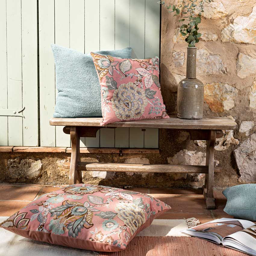Magma Kissen „Dunya“ mit floralem Muster in Rosa, Blau und Beige – stilvoll kombiniert mit Uni-Kissen auf rustikaler Holzbank in gemütlicher Outdoor-Atmosphäre.