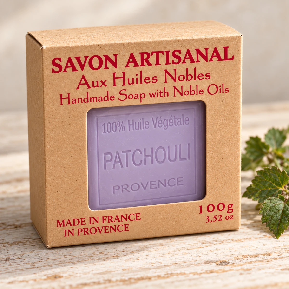 Maître Savonitto Seife Patchouli in vollständig sichtbarer Kraftkartonverpackung mit Sichtfenster, neben grünen Blättern auf hellem Holz.