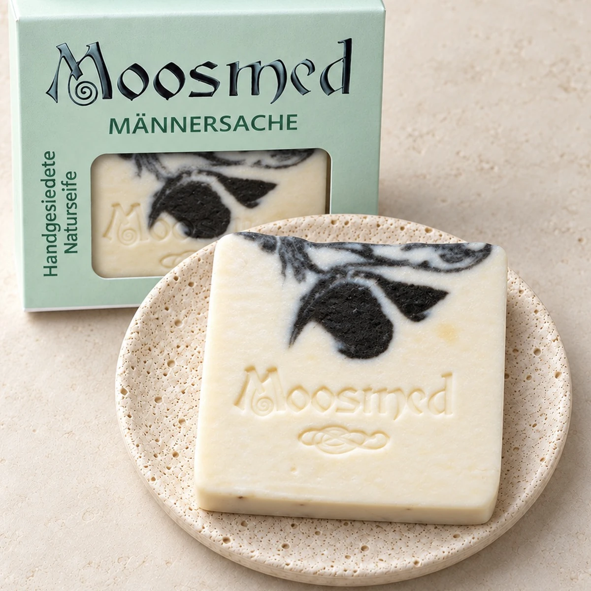 Cremefarbene Moosmed-Männersache-Seife mit schwarzer Marmorierung auf einem runden Travertinteller, dahinter die mintgrüne Verpackung.