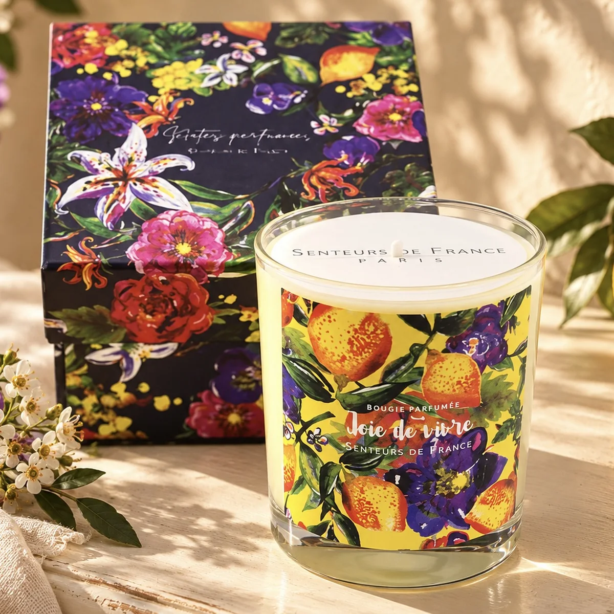 Duftkerze Joie de vivre von Senteurs de France mit gelbem Blütenetikett und floraler Geschenkbox auf hellem Holz mit Blüten und Zitrone.