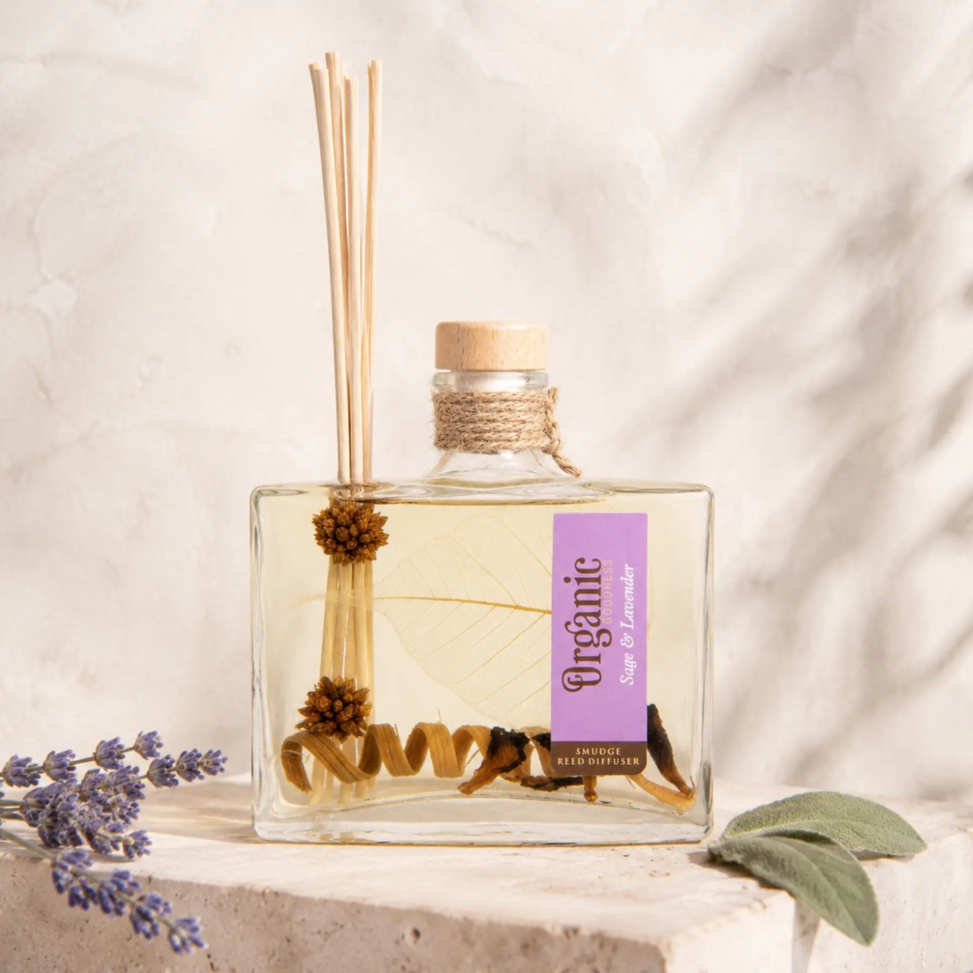 Organic Goodness Reed Diffuser Sage & Lavender mit Lavendelblüten und Salbeiblättern auf hellem Travertin.