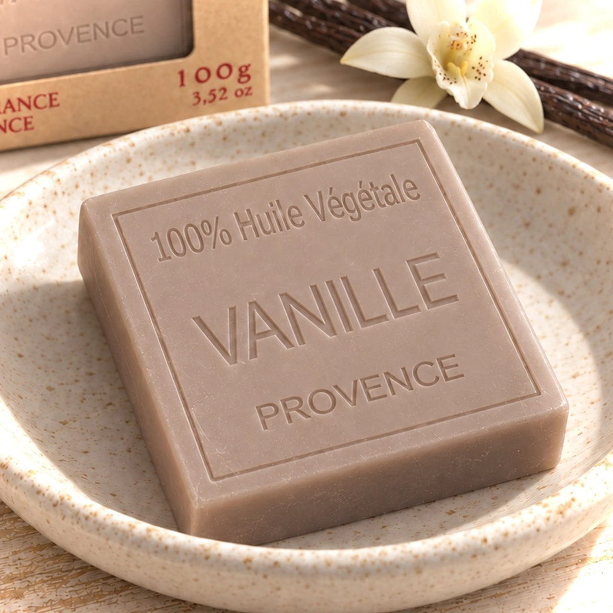 Maître Savonitto Vanilleseife auf einem hellen Keramikteller, mit Kartonverpackung dahinter und Vanilleschoten und einer hellen Blüte.