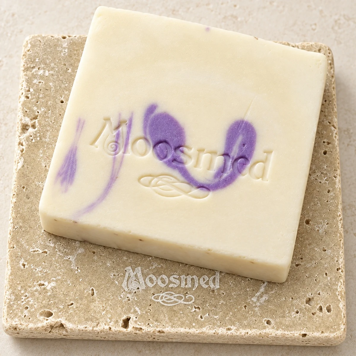 Cremefarbene Moosmed-Lavendel-Bergamotte-Seife mit violetter Marmorierung und geprägtem Logo auf einem quadratischen Travertinteller.