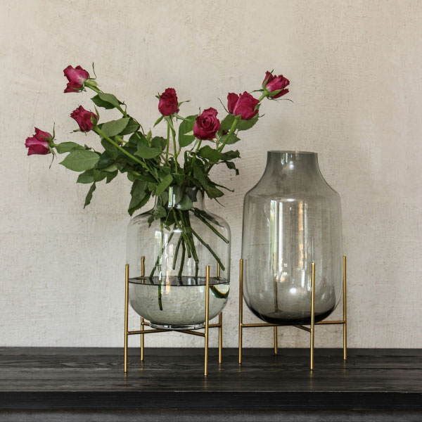 Glasvasen von Miljögården – elegante transparente Vasen in Rauchglas-Optik mit goldfarbenem Metallständer, stilvoll arrangiert mit Rosen auf dunkler Konsole.