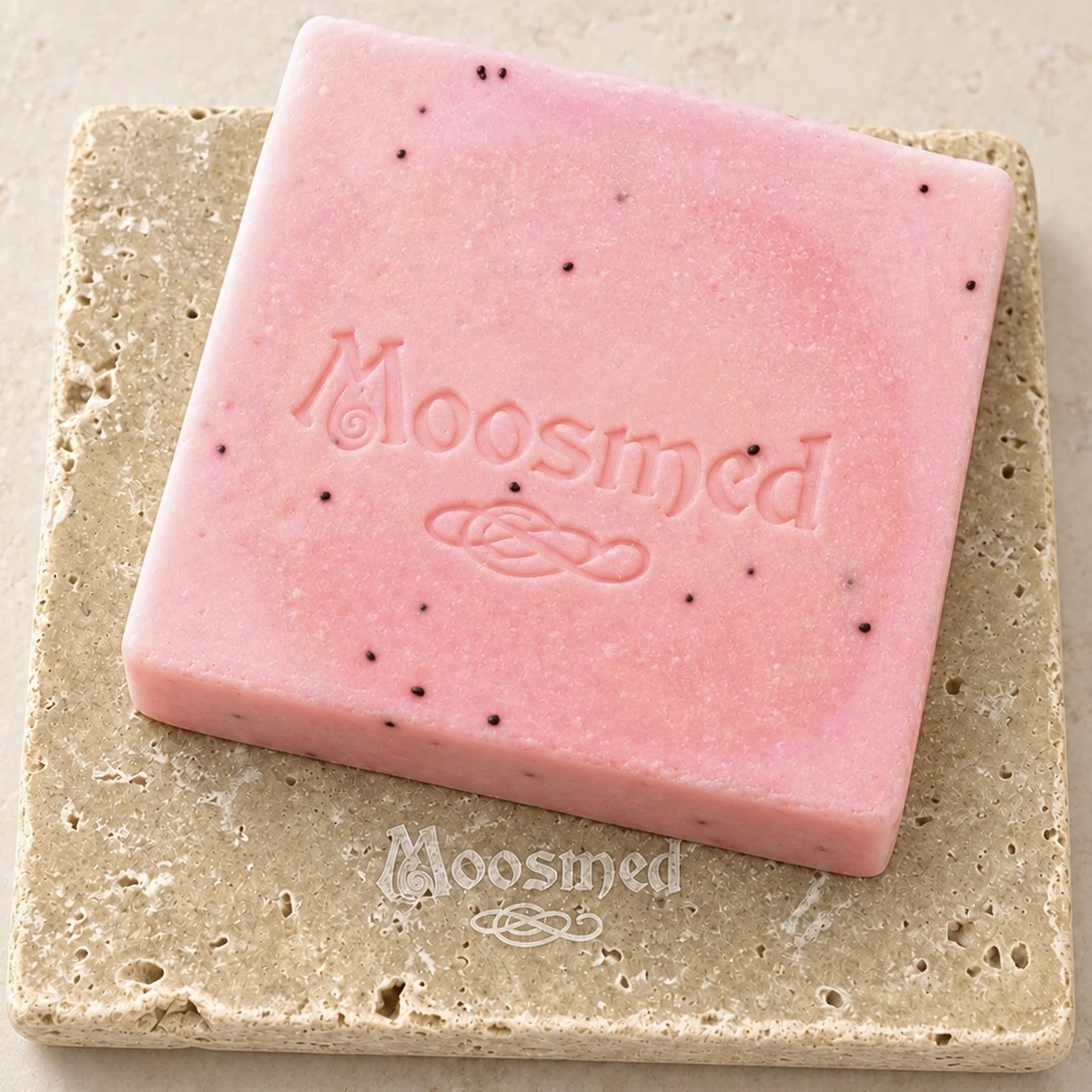 Rosafarbene Moosmed-Granatapfel-Seife mit dunklen Körnchen und geprägtem Logo auf einem quadratischen Travertinteller.